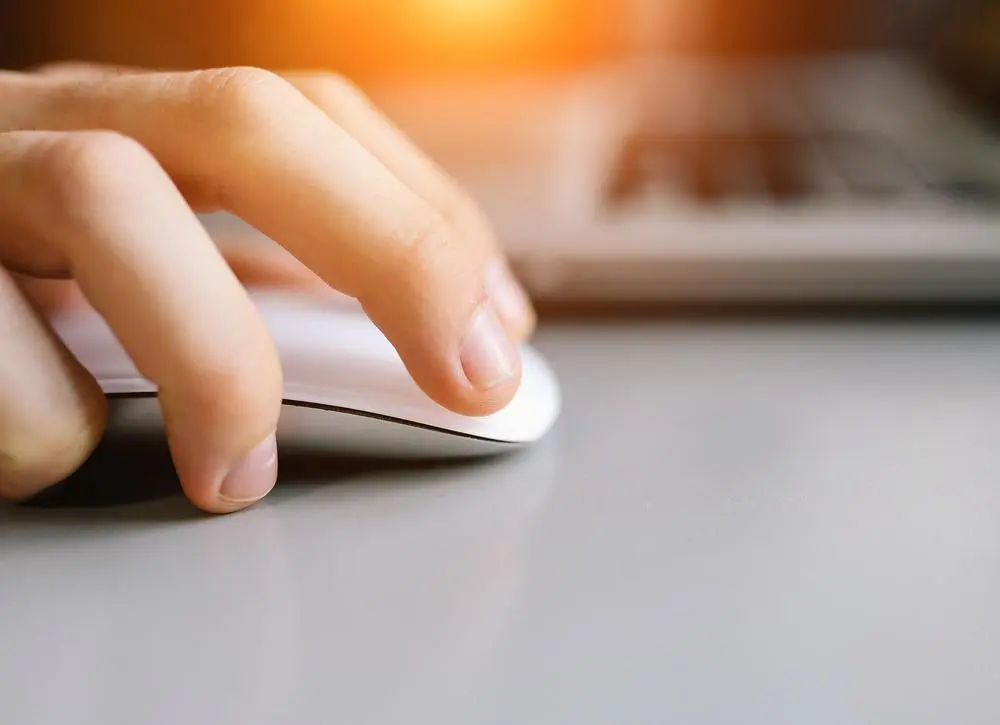 A hand using a white computer mouse on a gray surface, with a laptop in the background. Warm sunlight is softly illuminating the scene.