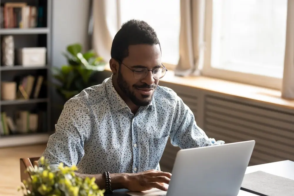 A person sits at a desk, smiling while using a laptop. They are wearing glasses and a patterned shirt. The room is bright, with a window and bookshelf in the background, and a plant is visible near the desk.