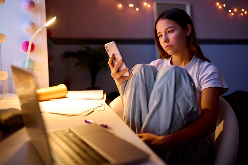 A teenage girl sits at a desk with a laptop, holding her phone and looking at it thoughtfully. She wears pajamas and is surrounded by study materials and warm string lights in a dimly lit room.