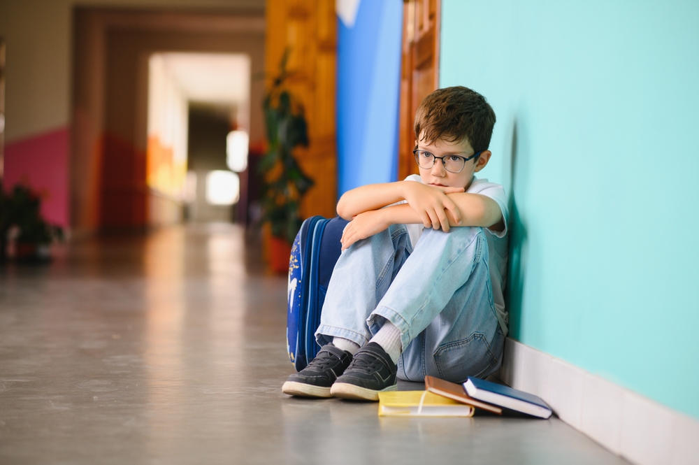A sad boy wearing glasses sits on the floor against a turquoise wall at school, hugging his knees with a backpack beside him and books lying on the ground.