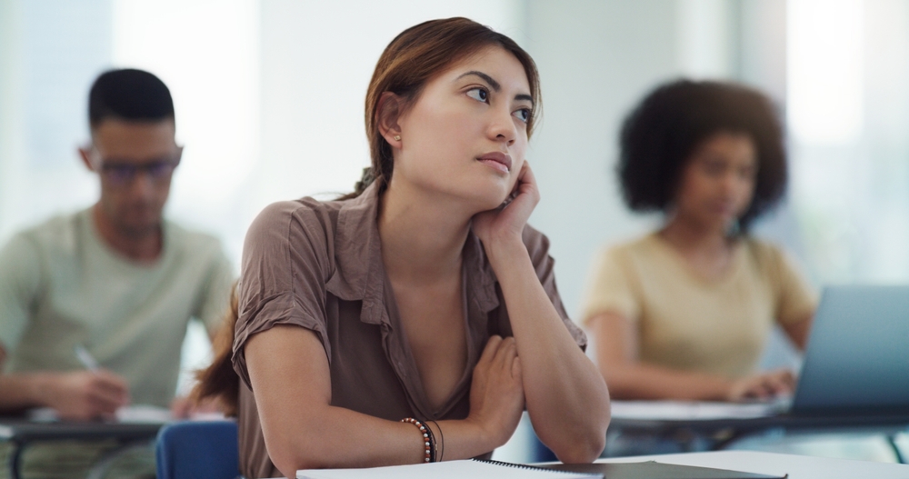 A young woman sits at a desk in a classroom, resting her chin on her hand and looking distracted or bored, while other students around her take notes or work on laptops.