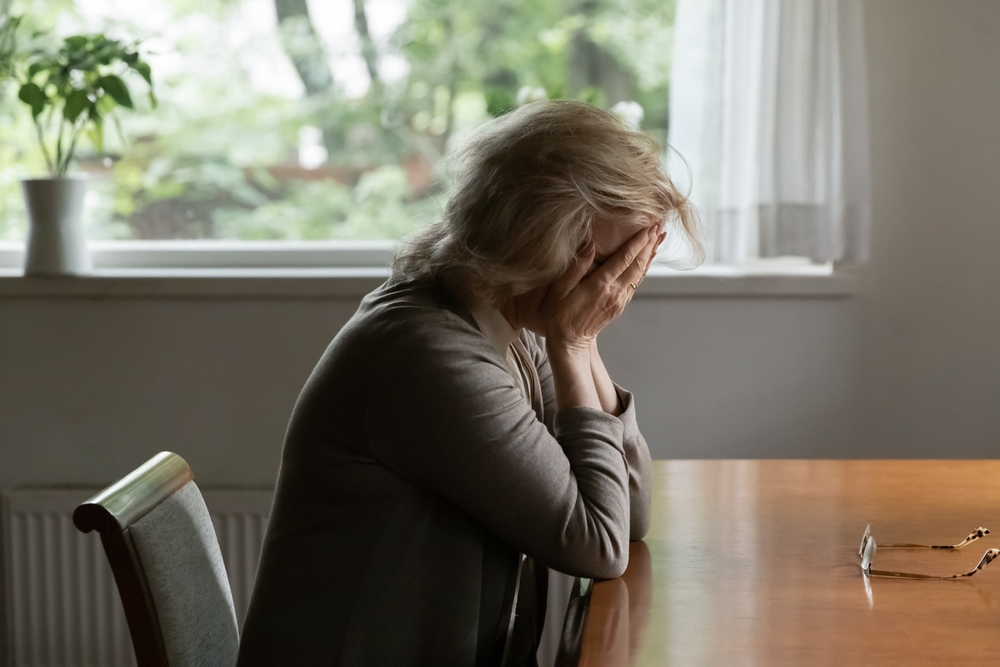An older woman sits at a table with her face in her hands, appearing distressed or upset. A pair of glasses lies on the table beside her. A window and a potted plant are visible in the background.