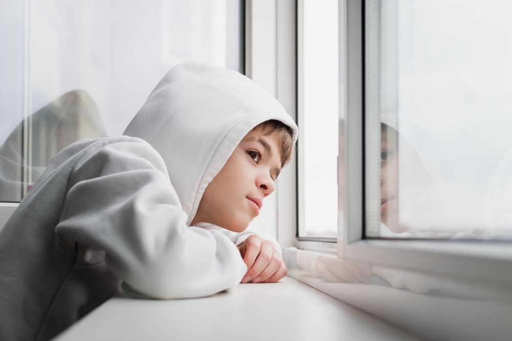 A young boy in a white hoodie leans on a windowsill, gazing thoughtfully out the window with his reflection visible in the glass.