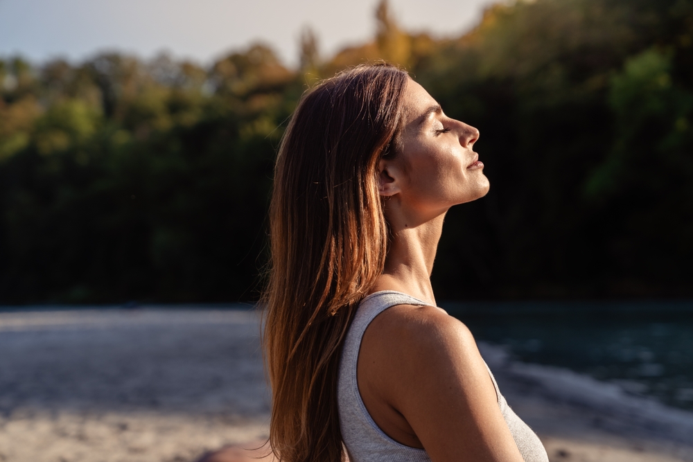 A woman with long hair stands outdoors near water, eyes closed, and faces the sunlight with a peaceful expression. Trees and a sandy shore are visible in the background.